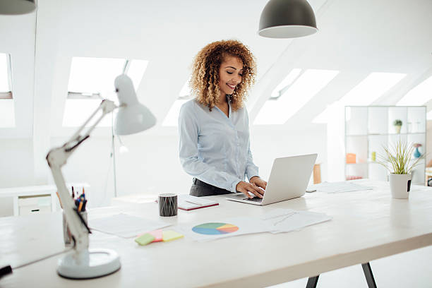 Latina Businesswoman Working In Her Office. Latina businesswoman working in her office. She is standing by the desk and using her laptop. Smiling. desk lamp photos stock pictures, royalty-free photos & images