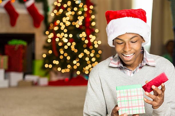 Handsome teenage or college age boy smiles happily while peeking inside of a Christmas gift. He is holding the gift while he opens up the lid. He is wearing a gray sweater and a santa hat. A Christmas tree, fireplace with stockings and gifts are in the background. Copy space is available.