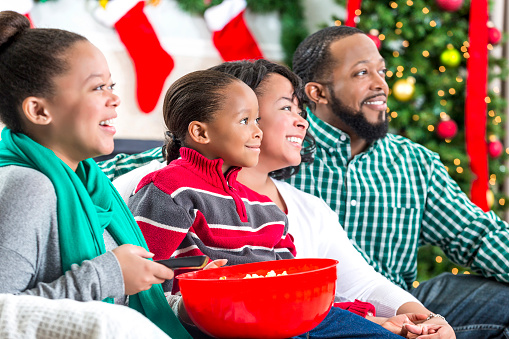 Happy African American family enjoys watching Christmas movie together while sitting on the cozy sofa. Mid adult parents and elementary age son and teenage daughter bond while watching the movie. The daughter is holding a red bowl full of popcorn and also holding the remote. A fireplace, stockings and lit Christmas tree are blurred in the background.