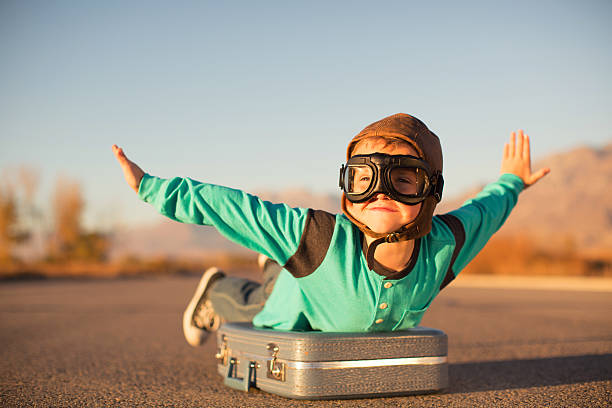 Young Boy with Goggles Imagines Flying on Suitcase A young boy with outstretched arms lies on top of a suitcase imagining he is flying on an airplane away and traveling to exotic locations. He is wearing a flight cap and goggles and has a large smile on his face. He loves to travel.  air vehicle photos stock pictures, royalty-free photos & images