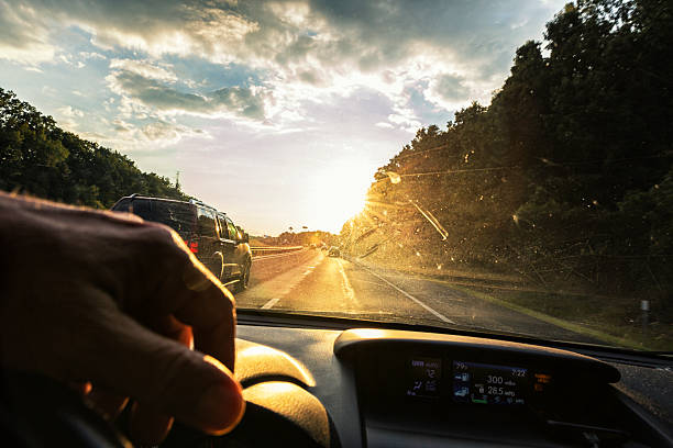 Driver with a hand on the steering wheel slightly tilted POV looking through the grubby, bug spotted, partially opaque, partially transparent speckled windshield of a car speeding next to another vehicle on an expressway following traffic heading directly into a bright late afternoon star-shaped sunburst sunset. Canon 5D Mark III.