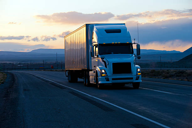 Modern semi truck trailer on twilight highway Huge modern big big semi truck with a trailer moving on the road in the evening twilight with headlights on the background of the picturesque hills with reflection burning rays of the evening sunset. truck mode of transport road transportation stock pictures, royalty-free photos & images