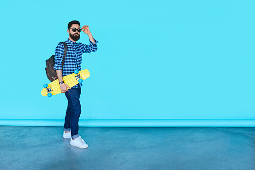 Full body portrait of young fashionable hipster man posing over blue background with copy space. Bearded male holding yellow skateboard and smiling
