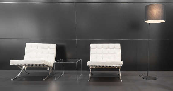 Brussels, Belgium - January 12, 2016: Pair of white modernist Barcelona design chairs and a modern lamp in front of a grey background. The Barcelona chair was designed by Ludwig Mies van der Rohe and Lilly Reich for the German pavilion for the International Exposition of 1929 in Barcelona. The chairs are on display at the 2016 Brussels motor show.