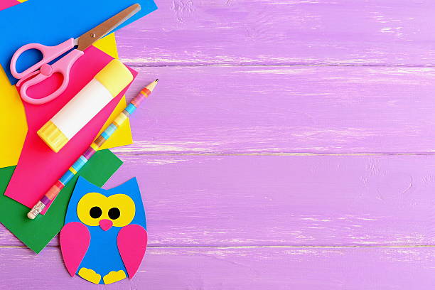 Coloured cardboard owl decor, scissors, glue stick, pencil, colored cardboard sheets on lilac wooden background with blank space for text. Children background