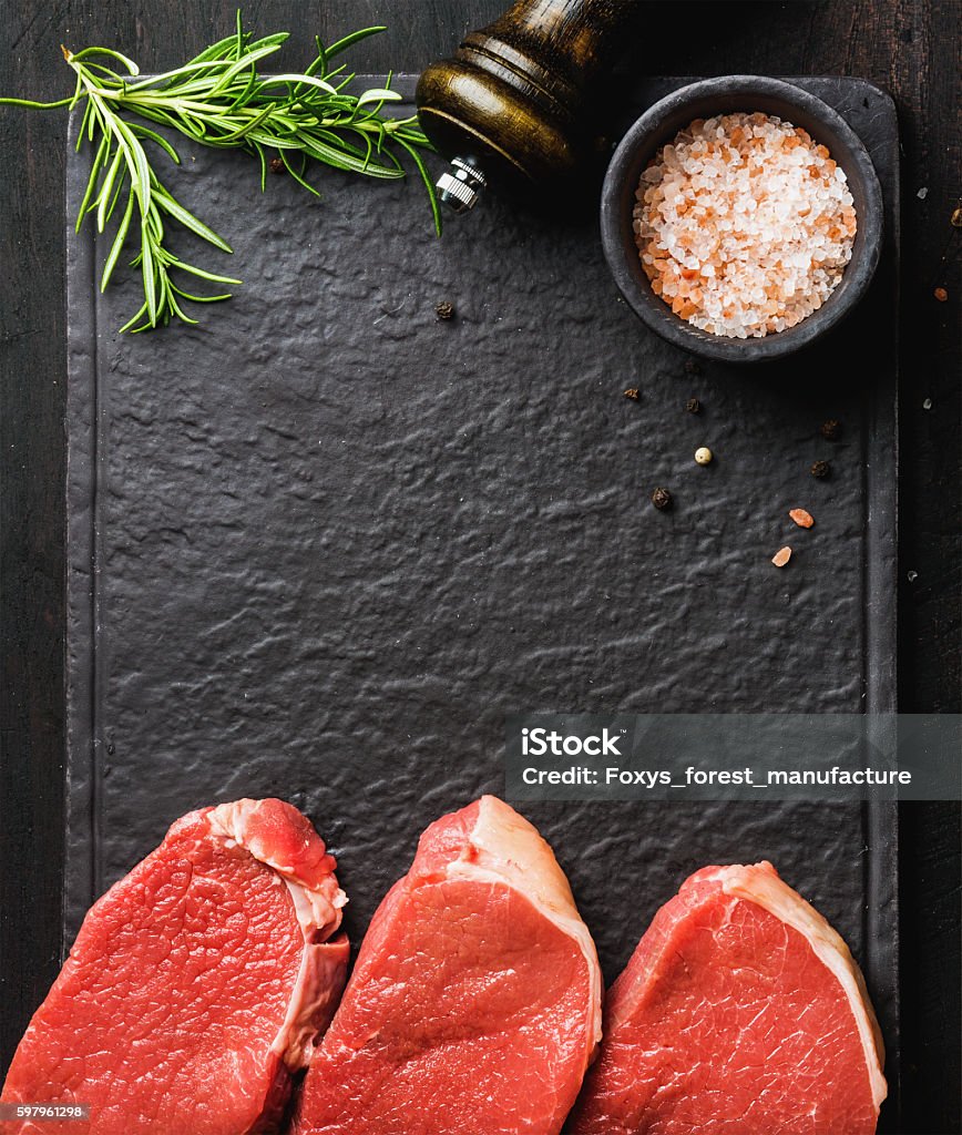 Raw beef Eye Round steaks with spices, rosemary. Copy space Raw beef Eye Round steaks with spices and rosemary on black slate stone board over dark wooden background, top view, copy space Healthy Eating Stock Photo