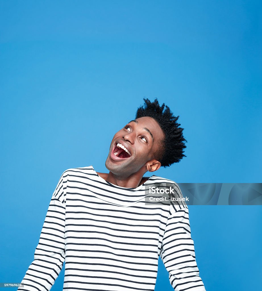 Excited afro american guy Portrait of happy afro american young man wearing striped top, looking up and laughing. Studio portrait, blue background. People Stock Photo
