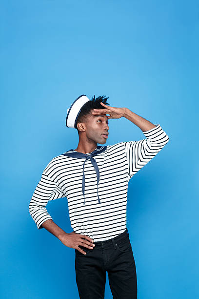 Portrait of afro american young man in sailor style outfit, looking away withe raised hand. Studio portrait, blue background.