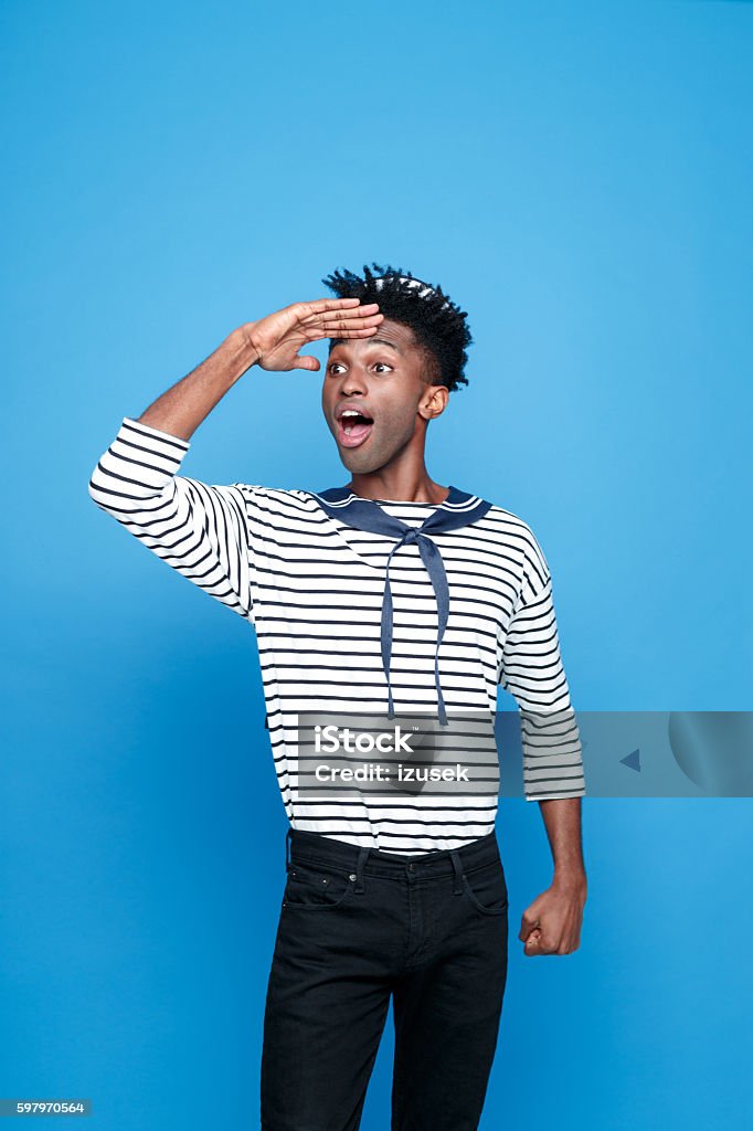 Excited afro american young sailor looking away Portrait of excited afro american young man in sailor style outfit, looking away withe raised hand and mouth open. Studio portrait, blue background. Sailor Suit Stock Photo