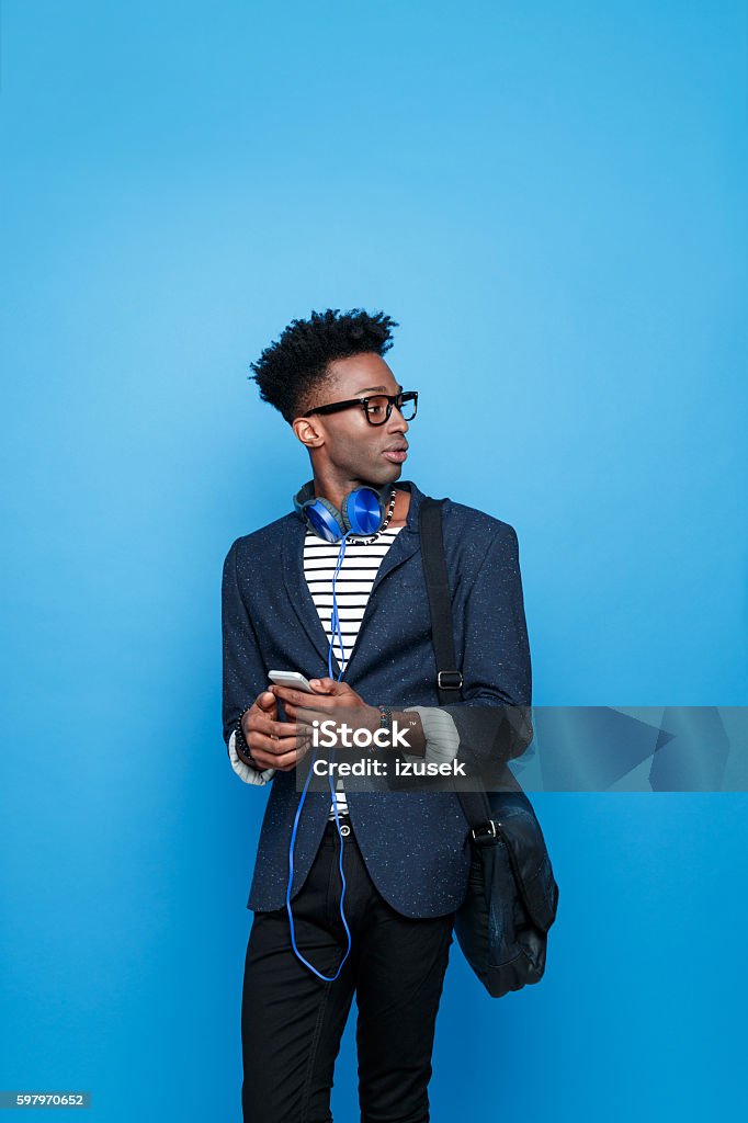 Afro american guy in fashionable outfit, holding smart phone Studio portrait of afro american young man wearing striped top, navy blue jacket, hat, nerd glasses and headphone, holding a mobile phone in hands. Studio portrait, blue background. Colored Background Stock Photo