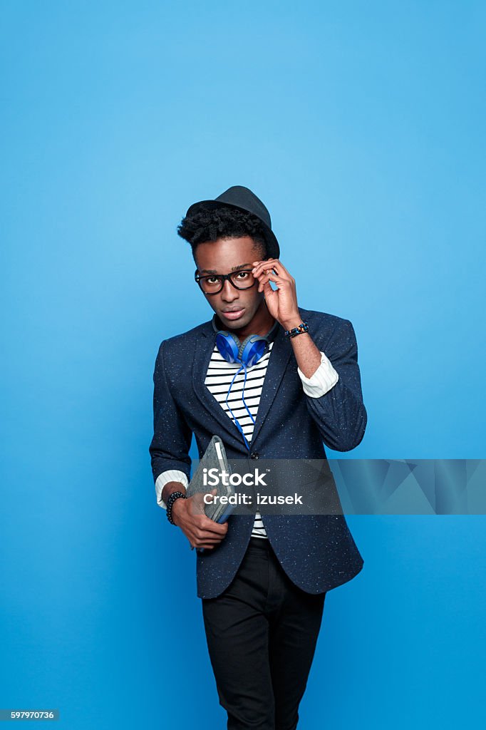 Afro american guy in fashionable outfit, holding notebook Studio portrait of afro american young man wearing striped top, navy blue jacket, hat, nerd glasses and headphone, looking at camera, holding a notebook in hand. Studio portrait, blue background. Adult Stock Photo