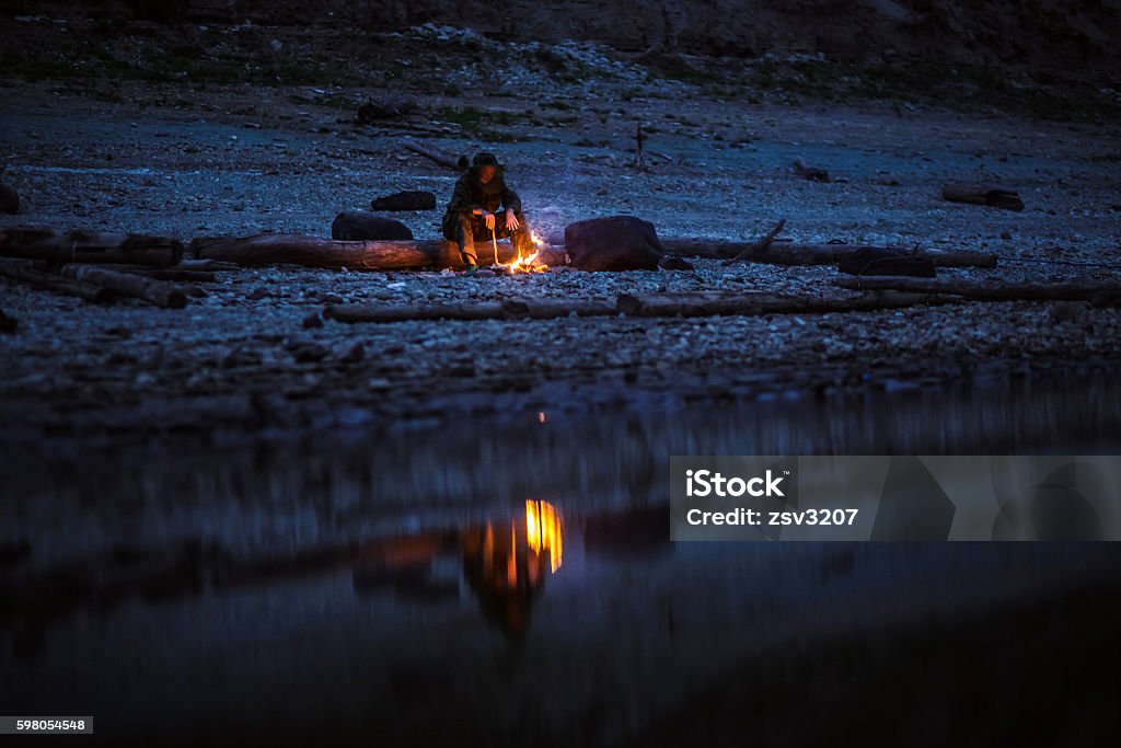 Hiker sitting near camping fire at river shore. Concept of Shot of a hiker at the dawn sitting near the camping fire. Concept of loneliness, choice, silence.High ISO, grainy image. Campfire Stock Photo