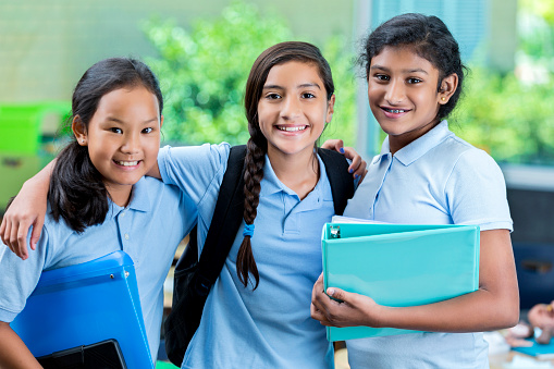 Three diverse middle school female students smile confidently on their first day of school. They have their arms around one another. They are holding notebooks and backpacks.