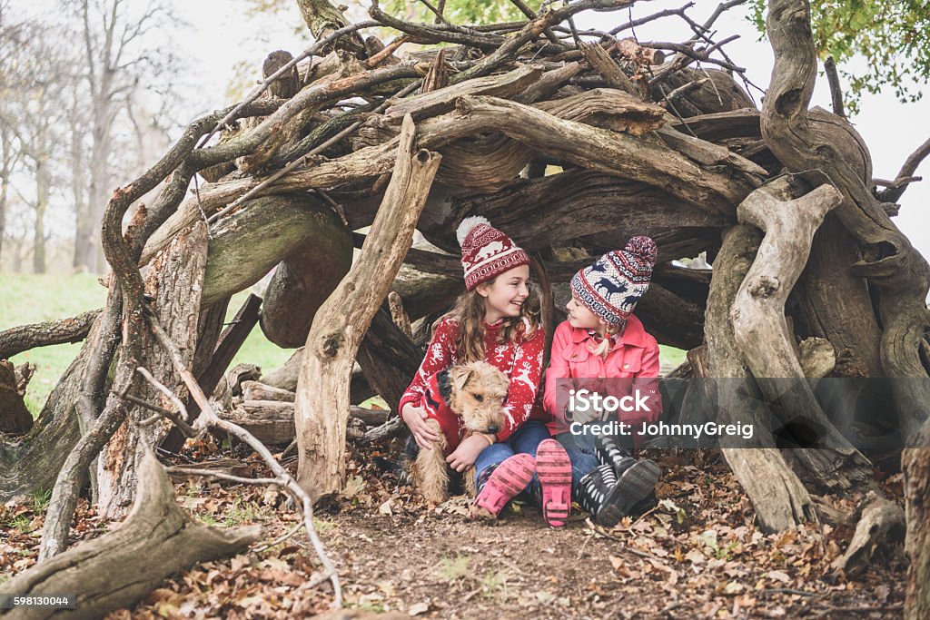 Two sisters in winter clothes in log den with dog Portrait of two female friends wearing wooly hats hiding in homemade log den Child Stock Photo