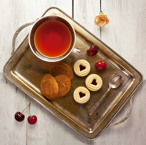 A photo of a vintage tray with a cup of tea, cookies, and cherries, shot from above on a light wooden boards texture; tea time concept
