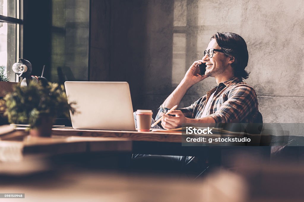 Staying in touch with his colleagues. Cheerful young man in casual wear looking through window and talking on the mobile phone while sitting near at the desk in creative office Men Stock Photo