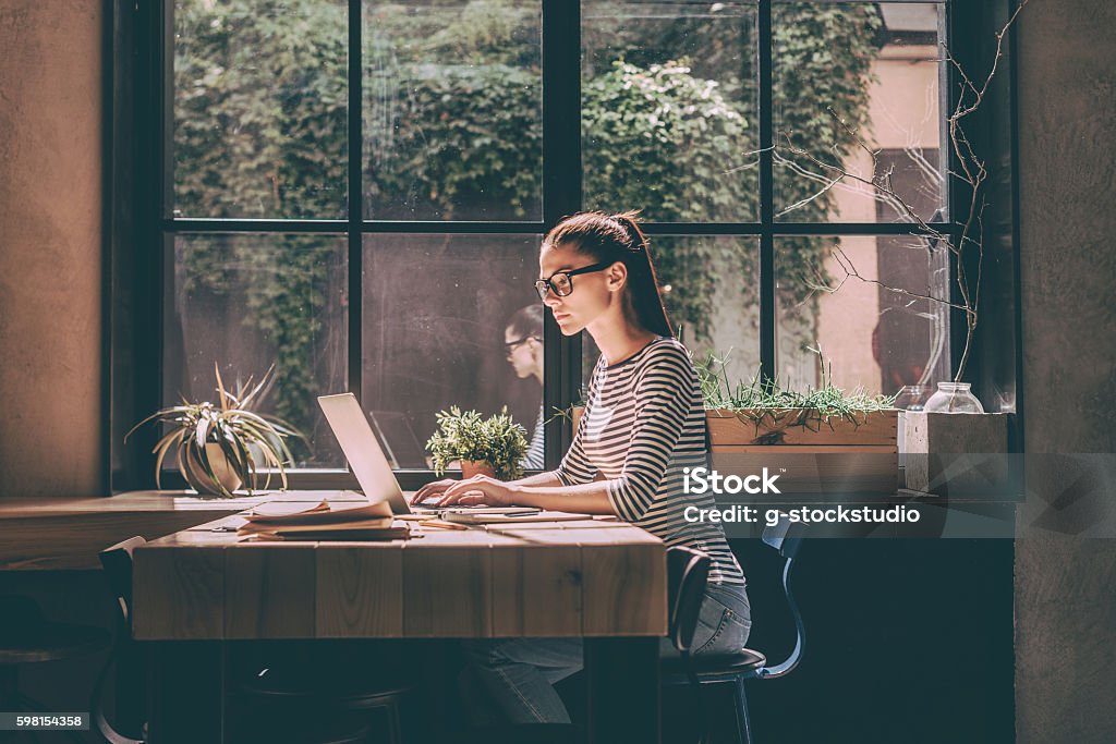 Concentrated at work. Confident young woman in smart casual wear working on laptop while sitting near window in creative office or cafe Women Stock Photo