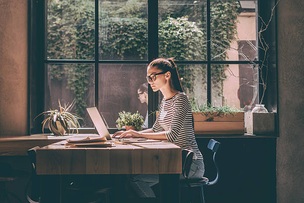 Concentrated at work. Confident young woman in smart casual wear working on laptop while sitting near window in creative office or cafe beautiful people office stock pictures, royalty-free photos & images