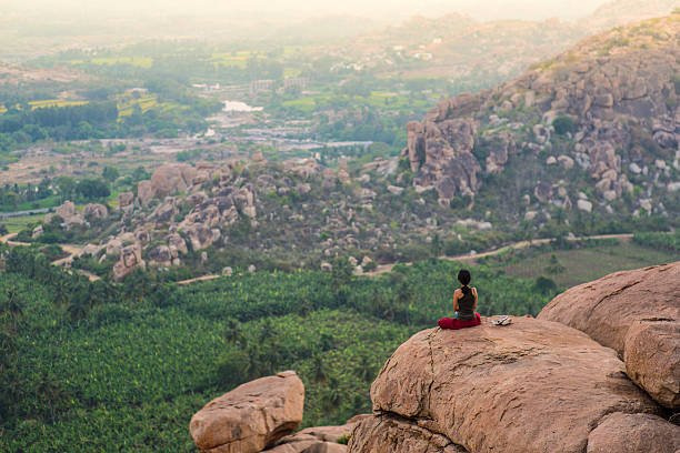 Hampi, India - March 22, 2015: A picture of a female tourist sitting in meditation on the top of Hanuman Temple mountain with a impressive view of the rocky landscape in front of her at sunset. Hampi is a popular tourist destination for backpackers in India.