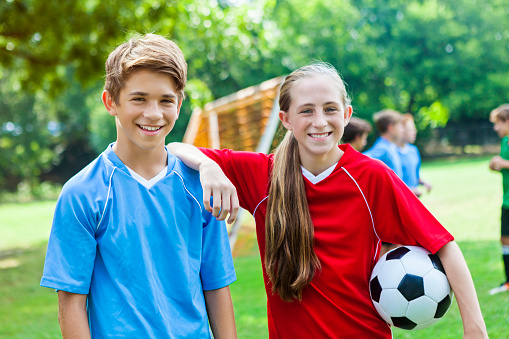 Teenage brother and sister or soccer friends kid around before soccer practice. The girl has her arm on the boy's shoulder and is holding a soccer ball. She has long blond hair in a ponytail across her shoulder. She is wearing a bright red jersey. The boy is wearing a blue jersey and has short brown hair. They are both smiling. Players are on the field in the background along with the soccer goal.