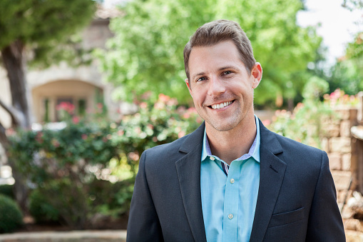 Young Caucasian business professional smiles While standing outdoors. He is wearing a jacket and a button-down shirt. He has brown hair.