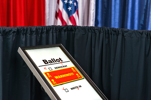 A hacked electronic voting machine, showing a red warning window with skull and cross bones icon at a polling station decorated in red, white and blue with an American flag in the background.