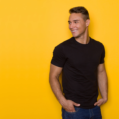 Smiling young man in black t-shirt and jeans, posing with hands in pockets and looking away over the shoulder. Waist up studio shot on yellow background.