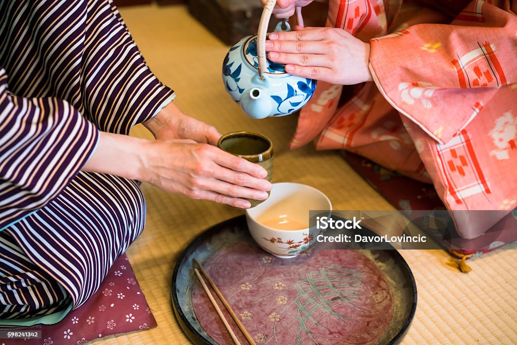 Dertail of having traditional Japanese tea in Kyoto Japan detail of two women in traditional kimono, kneeling on tatami preparing, pouring a  cup of tea which is inthe hands of one woman. They are in traditional Japanese old house on tatami. This is in Toei studios in Kyoto with old buildings from Samurai times. Kyoto City Stock Photo