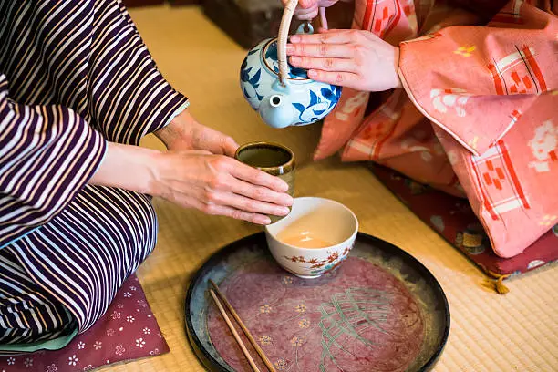 detail of two women in traditional kimono, kneeling on tatami preparing, pouring a  cup of tea which is inthe hands of one woman. They are in traditional Japanese old house on tatami. This is in Toei studios in Kyoto with old buildings from Samurai times.