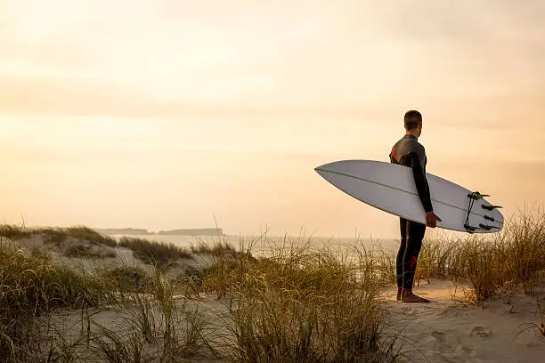 A surfer with his surfboard at the dunes looking to the waves