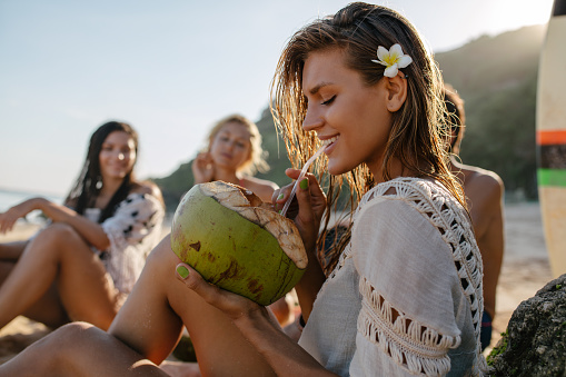 Side portrait of attractive young woman drinking coconut juice with her friends sitting in background. Young people relaxing on the beach on summer vacation.
