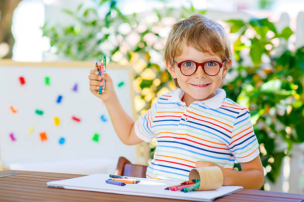 Funny adorable little kid boy with glasses holding wax crayons pens. Happy child and student is back to school and looking at pupil's stuff on warm day.