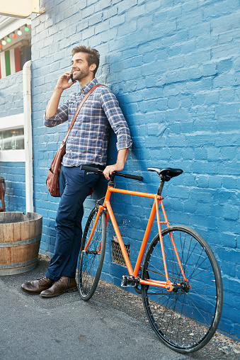 Shot of a handsome young man leaning against a wall with his bike talking on a cellphone