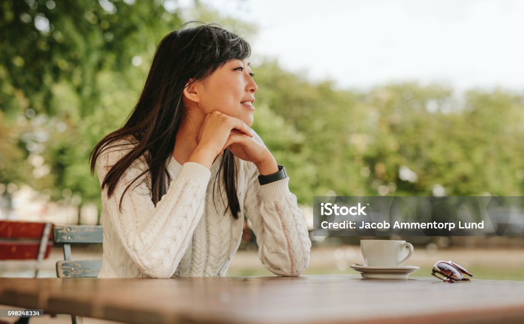 Asian woman spending free time at outdoor cafe Happy young woman sitting at cafe with a cup of coffee on table and looking away. Chinese female spending free time at outdoor cafe. Asian and Indian Ethnicities Stock Photo