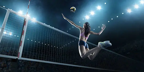 Beautiful female volleyball player performs an emotional game moment on the indoor volleyball stadium with bleachers full of people. She is wearing an unbranded sports cloth.
