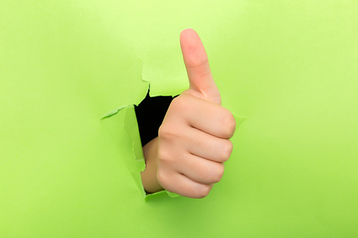 Thumbs up through the paper hole isolated on green background