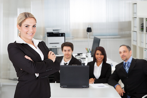 Portrait of young businesswoman standing arms crossed while colleagues sitting at desk in office