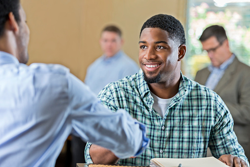 Young African American man smiles while shaking hands with a businessman. The young man is interviewing for a position. He is wearing a plaid shirt and has short black hair and facial hair. People are in the background.