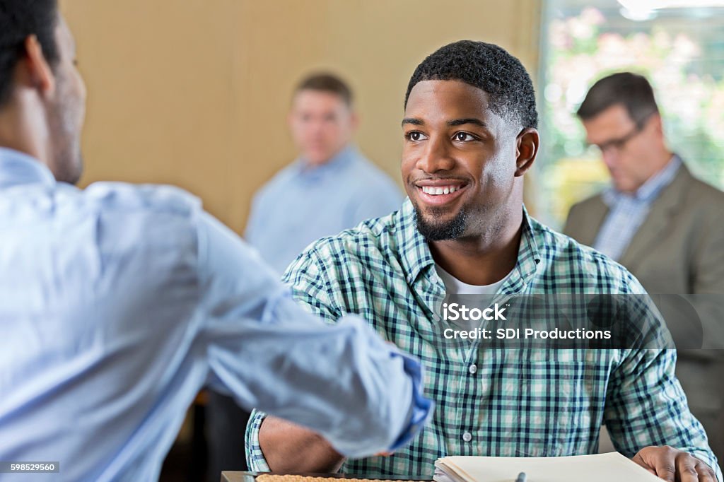 Handsome young African American man at job interview Young African American man smiles while shaking hands with a businessman. The young man is interviewing for a position. He is wearing a plaid shirt and has short black hair and facial hair. People are in the background. Job Fair Stock Photo