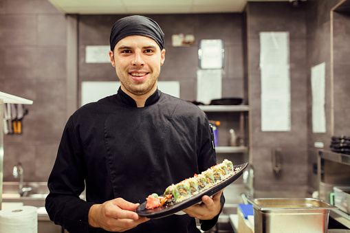 Chef posing in restaurant kitchen with meal on plate prepared for serving. He made dragon maki sushi.