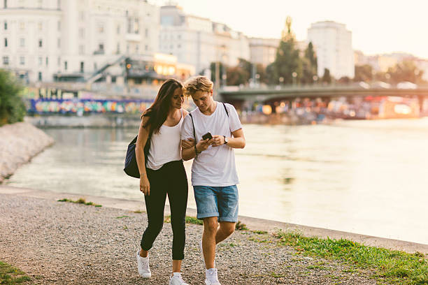 Teenage Couple Mobile Gaming Outdoors. Teenage couple walking near Danube river in Vienna at sunset. Boy holding smart phone and they are mobile gaming. Cityscape in background. Both are austrian models, couple in real life. teenage girls dusk city urban scene stock pictures, royalty-free photos & images