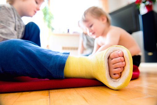 Close up of broken leg in cast of a little boy playing with his little brother sitting on the wooden floor. Childrens daytime fun. Happy to be at home.