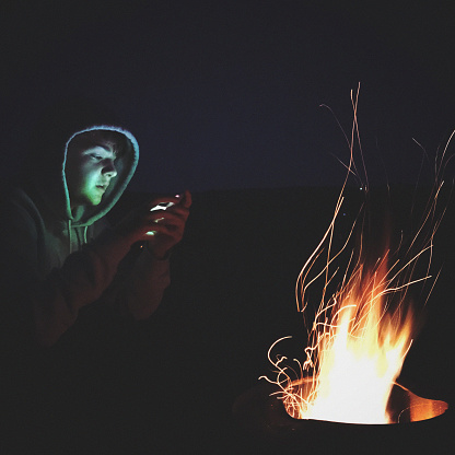 Teenager boy using smartphone by the campfire outside the tent at the campsite in Somerset, England