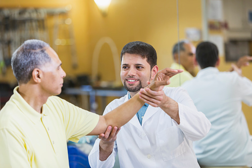 An Hispanic physical therapist or orthopedic doctor wearing a white lab coat, helping a male mixed race African American patient who has injured his arm.