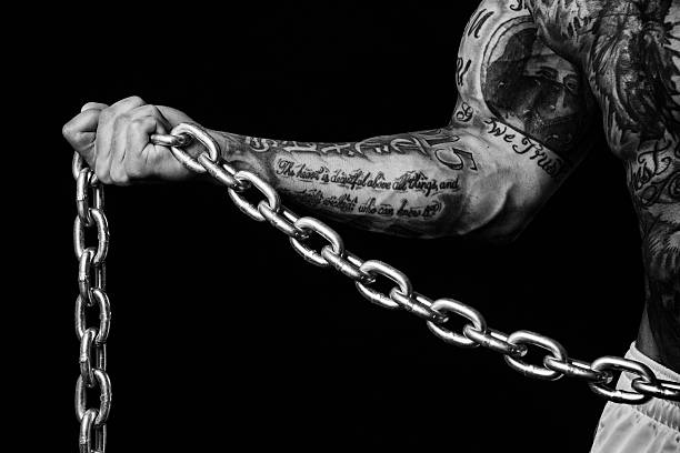 Isolated Arm and Hand Holding a Chain A tattooed muscular arm and hand holding a thick chain against a black background. forearm tattoos men stock pictures, royalty-free photos & images