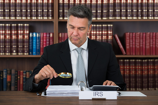 Mature male tax auditor examining documents with magnifying glass at table in office