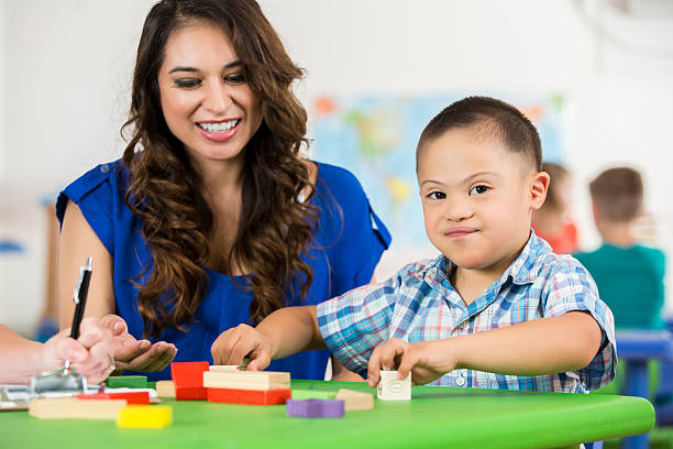 Hispanic Down Syndrome child playing with blocks at daycare Hispanic Down Syndrome boy looking at camera while playing with blocks at his daycare. He and his Hispanic female teacher are sitting at a table working together. special education stock pictures, royalty-free photos & images