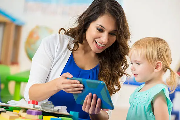 Smiling Hispanic female teacher shows a digital tablet to an interested blonde Caucasian little girl. They are sitting at a table together in a daycare learning center looking at something on the tablet.