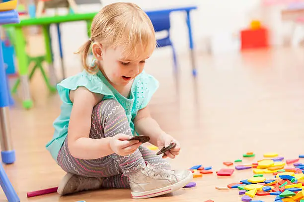 Cute Caucasian female preschooler playing with toys while sitting on the floor in her daycare. Blonde toddler with pigtails plays with puzzle pieces on the floor in the learning center.