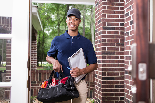 African descent repairman or blue collar/service industry worker make service/house call at customer's front door. He holds his clipboard and tool box filled with work tools.  Inspector, exterminator, electrician.  Red brick home.